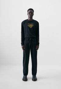 Chemise noire à manches longues avec du texte doré et des détails graphiques, assortie à un pantalon noir et des chaussures noires. Design simple et minimaliste.