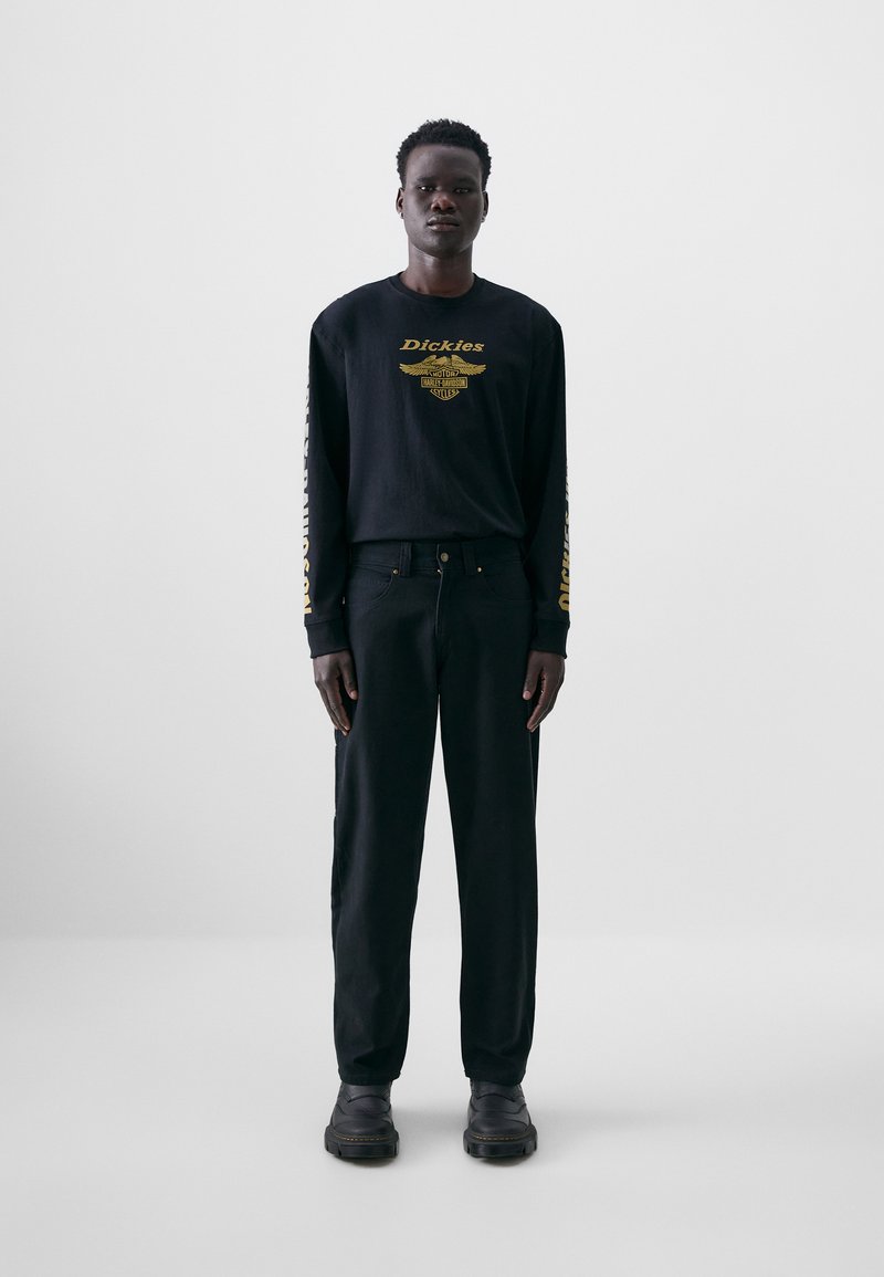 Chemise noire à manches longues avec du texte doré et des détails graphiques, assortie à un pantalon noir et des chaussures noires. Design simple et minimaliste.