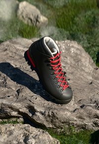 Schwarzer Wanderstiefel mit roten Schnürsenkeln auf einer felsigen Oberfläche, umgeben von Gras, das die strapazierfähige Sohle und den gepolsterten Knöchelkragen hervorhebt.