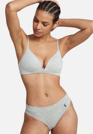 Mujer con el cabello trenzado posa vistiendo un sujetador gris claro y ropa interior a juego con un pequeño logo azul marino en la cadera, sobre un fondo blanco.