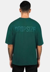 Grünes Baumwoll-T-Shirt mit kurzen Ärmeln, Rundhalsausschnitt und dem Wort "DROPSIZE", das groß und farblich passend auf dem Rücken aufgedruckt ist.