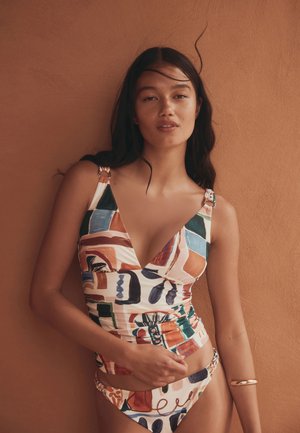 Femme aux longs cheveux foncés portant un maillot de bain coloré à motifs abstraits, debout contre un mur couleur terre cuite.