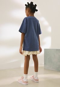 T-shirt bleu surdimensionné, superposé avec une jupe beige volantée. Baskets Adidas roses avec des accents blancs et des chaussettes blanches côtelées.