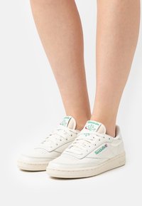 White Reebok sneakers with green and red logo worn on bare legs against a plain white background.