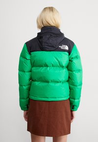 Zelena i crna puffer jakna s kapuljačom na zatvarač, kvadriranom dizajnom i logotipom na gornjem dijelu leđa, u kombinaciji s smešnom suknjom.