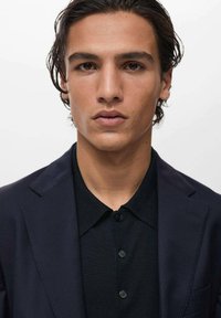 Ciemnoniebieska marynarka na czarną koszulę z kołnierzykiem, o prostym designie i gładkiej fakturze. Skupienie na wyrazistych, symetrycznych rysach twarzy.