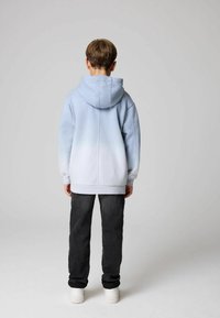 Ljusblå gradiente hoodie med en rymlig huva, ribbade cuffs och nederkant, i kombination med mörka jeans och vita sneakers. Sedd bakifrån.