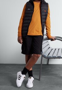 Czarna pikowana kamizelka na pomarańczowej koszuli z długim rękawem, do tego czarne szorty, białe sneakersy z czarnymi paskami i czarne skarpetki.