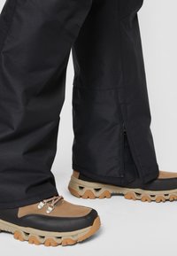 Zwarte waterdichte broek met ritsen aan de onderkant, gecombineerd met bruine en zwarte wandelschoenen met gestructureerde zolen en veters.