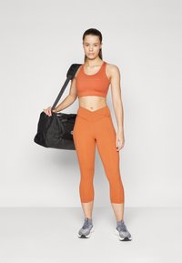 Idrettsutøver i en oransje sports-BH og matchende leggings, som holder en svart treningstaske. Leggingsene har høy midje og sømløst design.