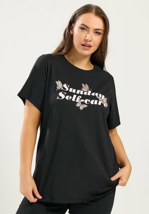 Kvinde med langt brunt hår iført en sort t-shirt med teksten "Sunday Selfcare" og sommerfuglegrafik på forsiden.
