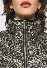 Doudoune avec un motif léopard beige et noir, présentant un col haut, des poches zippées et des accents en métal doré.