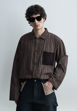 Mężczyzna z kręconymi włosami, noszący czarne okulary przeciwsłoneczne, brązową koszulę w paski z czarną teksturowaną kieszenią oraz ciemne spodnie z beżowymi kieszeniami w kratę.