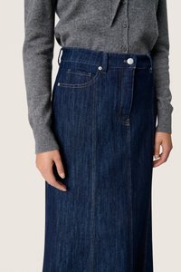 Sötétkék farmer szoknya magas derékkal, oldalsó zsebekkel és látható varrásokkal. Szürke pulóverrel viselve, amely kiemeli a szabott fazont.