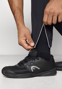 Melnas sporta kedas ar acs materiālu un gumijas zoli, ar pelēku logo. Rokas atver melnas bikses ar redzamu apdari.