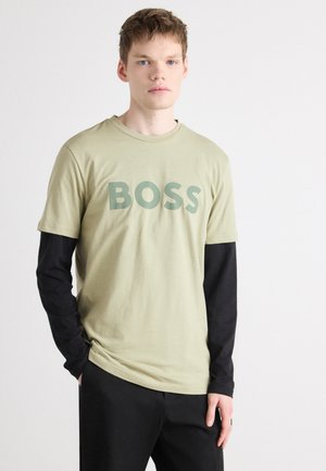 Olivenfarbene T-Shirts mit kurzen Ärmeln, auf denen in dunklerem Grün das große Wort "BOSS" zu sehen ist. Über einem schwarzen Langarmhemd getragen, mit lockerer Passform.