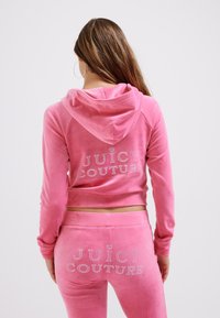 Rozā velūra treniņtērps ar īsu kapuci un atbilstošām biksēm. Aizmugurē ir "JUICY COUTURE" logo ar kristāliem.