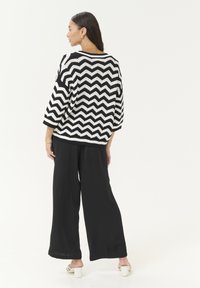 Pull en tricot noir et blanc à motif chevron avec des manches trois-quarts, associé à un pantalon large noir et des sandales à talons blancs.