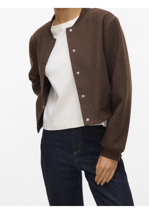 Persona con una chaqueta marrón abotonada sobre una camisa blanca y jeans azul oscuro, de pie con una mano relajada a un lado.