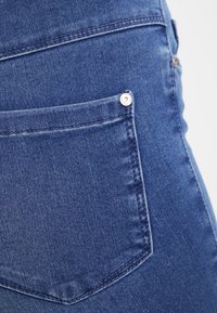 Denim jeans featuring a back pocket with a silver stud and visible stitching. The fabric is blue with a soft texture and slight fading.