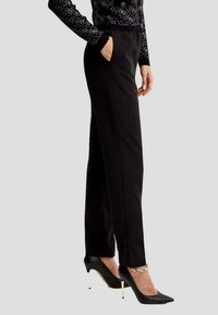Pantaloni neri su misura con tasche anteriori, taglio slim e texture liscia, abbinati a tacchi neri a punta con dettagli dorati.