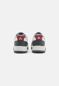 Sneakers low-top con una toma superiore in sintetico bianco, dettagli in rosso e grigio, superfici testurizzate e una suola in gomma nera solida.