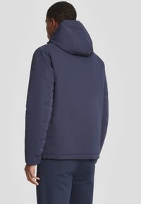 Giacca imbottita blu navy con cappuccio, caratterizzata da una texture liscia e un design semplice; include un patch con logo sulla spalla.