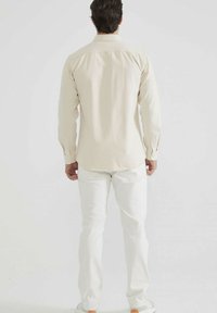 Un homme se tient de dos, portant une chemise beige à manches longues, un pantalon blanc et des chaussures blanches, devant un fond clair.