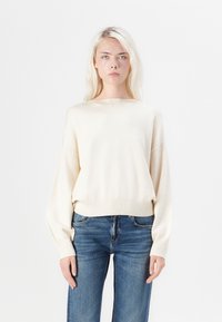Crème kleurige gebreide trui met een ruime halslijn en verlaagde schoudernaad, gecombineerd met hoge taille blauwe jeans. Zachte textuur en een relaxte pasvorm.