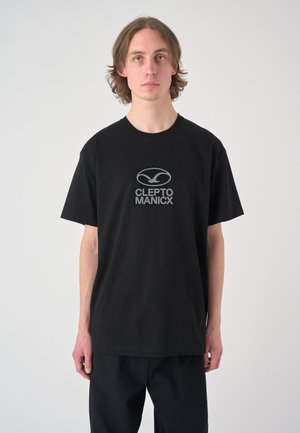 Junger Mann mit schulterlangen Haaren, der ein schwarzes Cleptomanicx T-Shirt und schwarze Hose trägt, steht vor einem schlichten hellen Hintergrund.