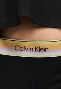 Κοντινό της κάτω πλάτης ενός ατόμου που δείχνει μαύρο παντελόνι με ουράνιο τόξο σε βαθμιδωτή μετάβαση στη μέση Calvin Klein και μέρος μαύρου τοπ.