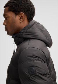 Giacca imbottita grigia con cappuccio, caratterizzata da una superficie testurizzata e da una distintiva toppa nera sulla manica sinistra. Presente cordoncino regolabile.