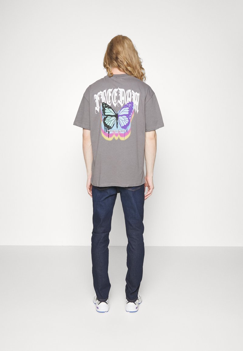 Szara koszulka z kolorową grafiką motyla oraz napisem "WOLNOŚĆ" w stylizowanej czcionce na plecach; zestawiona z ciemnymi dżinsami.