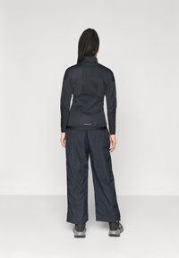 Črna tehnična jakna z visokim ovratnikom, v kombinaciji z ohlapnimi črnimi hlačami. Očitne šive in teksturirana tkanina na obeh kosih.