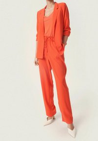 Pomarańczowa marynarka z dopasowanymi spodniami na sznurku. Miękka tkanina, luźny krój i stylowe białe obcasy dopełniają całość.
