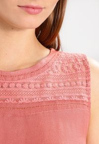 Kortärmad topp i mjuk rosa tyg, med spetsdetaljer och broderade mönster längs halsringningen. Slätt, texturerat material.