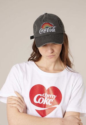 Personne portant une casquette noire Coca-Cola avec un logo arc-en-ciel et un T-shirt blanc avec un cœur rouge et le texte « cherry Coke », les bras croisés, regardant vers le bas.
