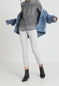 Chaqueta vaquera (azul claro), camiseta gris con bolsillos en el pecho, vaqueros pitillo gris claro con cremalleras laterales, y botines negros.