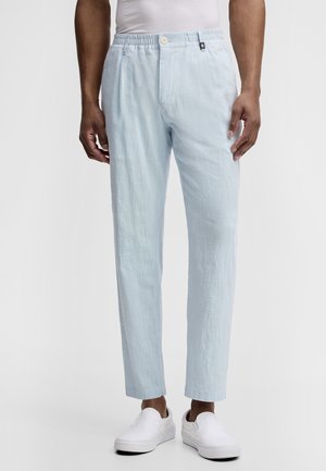 Man draagt lichtblauwe linnen broek met elastische tailleband en knoopsluiting, gecombineerd met witte instapschoenen en een wit overhemd.