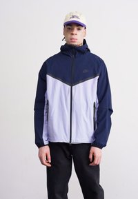 Veste Nike bleu marine et violet clair avec fermeture éclair sur le devant, capuche et poches latérales. Présente un accent noir sur la poitrine. Texture lisse.