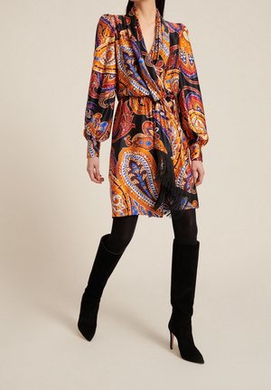 Kobieta ubrana w sukienkę do kolan z długim rękawem w wzór paisley w kolorach pomarańczowym, niebieskim i czarnym, do tego czarne rajstopy i czarne, sięgające do kolan botki na obcasie.