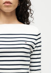 Γυναίκα με σγουρά μαλλιά που φοράει άσπρο πουκάμισο με οριζόντιες μπλε ναυτικές ρίγες και μικρό λογότυπο στο στήθος.