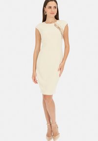 Beżowa dopasowana sukienka z krótkimi rękawami i koronkowymi detalami na lewym ramieniu. Gładka tekstura, do kolan, zestawiona z cielistymi szpilkami na kostce.