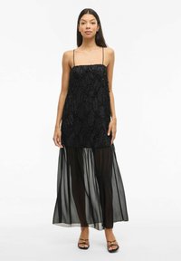 Juoda suknelė su tekstūruotu kūnu ir plonomis petnešėlėmis; sluoksniuota, permatoma sijonas. Yra sudėtingų audinio detalių ir sklandus siluetas.