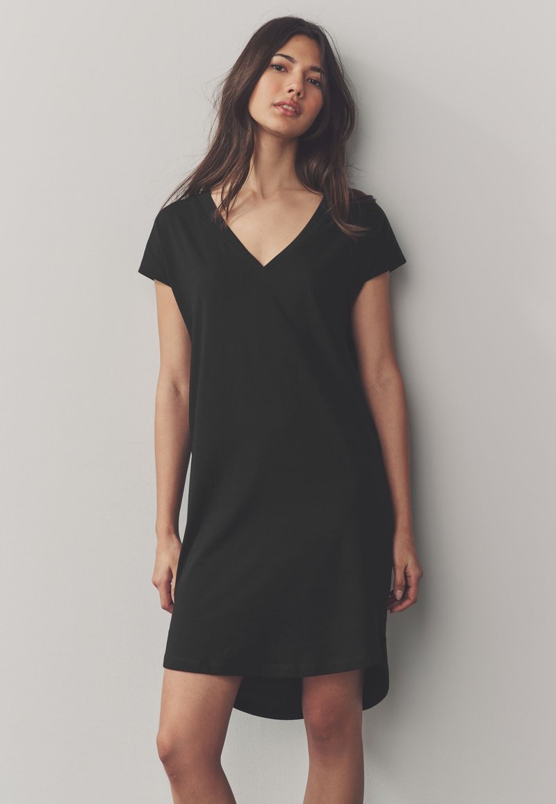 Femme aux longs cheveux foncés portant une robe noire à manches courtes avec un col en V, debout contre un mur gris clair.