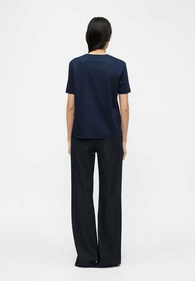 Γυναίκα με μακριά σκουρόχρωμα μαλλιά που φοράει μπλε κοντομάνικο μπλουζάκι και χαλαρά μαύρα παντελόνια, στεκόμενη με την πλάτη προς την κάμερα.