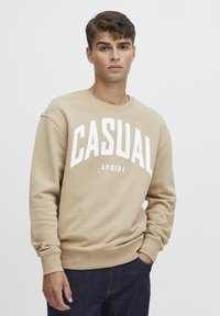 Béžová mikina s kulatým výstřihem a b�ílým nápisem „CASUAL FRIDAY“, se žebrovanými manžetami a spodním lemem, vyrobená z jemné, strukturované látky.