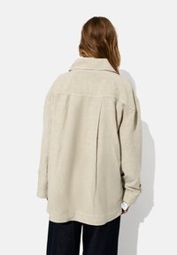Beige linned skjortejakke med krave og drop shoulders, prydet med en rygfold og opsmøgede ærmer. Fin tekstur, løs pasform.