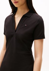 Schwarzes Polohemd mit V-Ausschnitt und kurzen Ärmeln. Mit einem kleinen Farblogo auf der oberen linken Seite. Hergestellt aus einem glatten, atmungsaktiven Stoff.