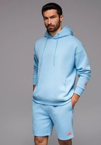Lichtblauwe sweatshirt met een capuchon en een vozak, gecombineerd met bijpassende shorts. De stof voelt zacht aan, met ribgebreide manchetten en logo-accenten.